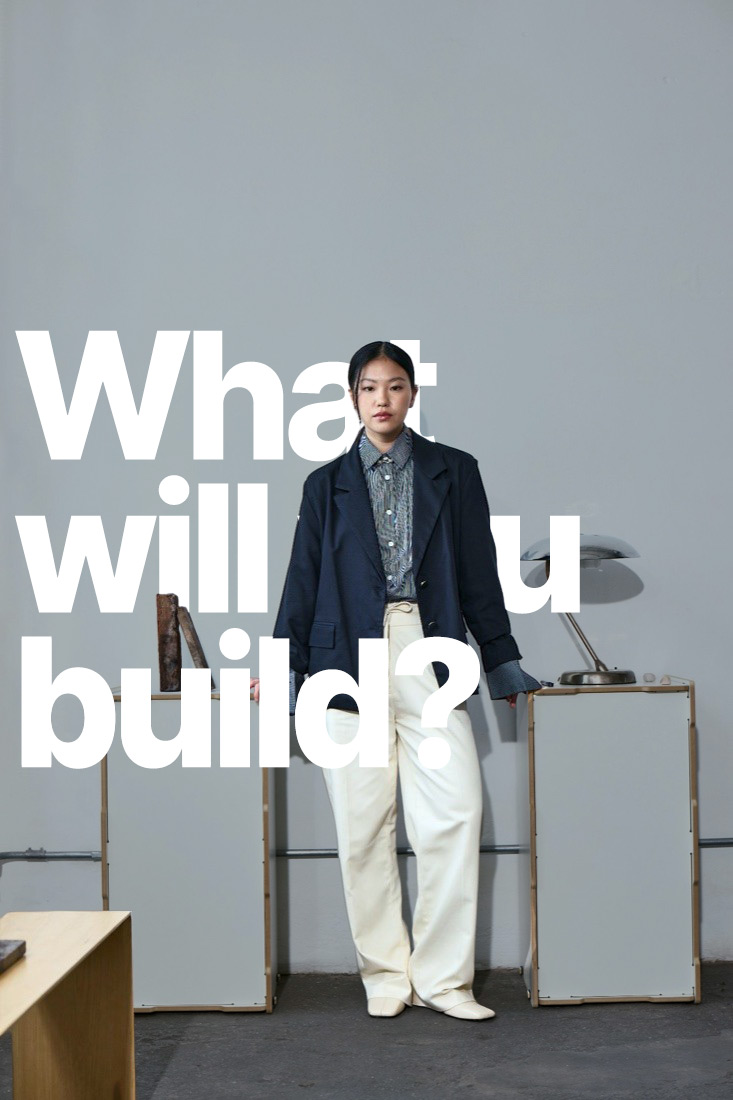 A woman in a navy blazer, striped shirt and cream trousers between two white plinths and a chrome lamp
