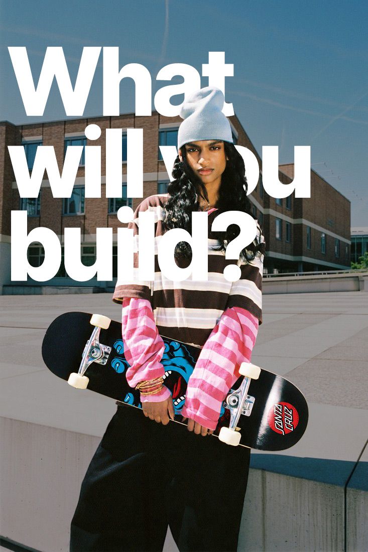 A woman in a grey beanie and striped rugby shirt holding a skateboard on a campus plaza